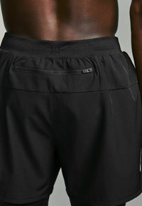 Shorts de sport noirs avec une texture lisse, une taille élastique et une poche zippée à l'arrière pour le rangement. Présente un design épuré et minimaliste.