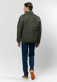 Tmavo zelená nafukovacia bunda s horizontálne prešívanou textúrou, stojatým golierom a elastickými manžetami, nosená cez modré džínsy a svetlé topánky.