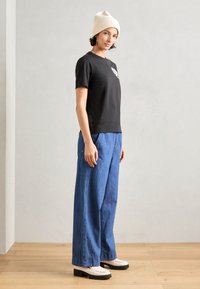 T-shirt nera a maniche corte con design grafico, abbinata a pantaloni larghi in denim blu e scarpe con platform color crema. Il modello indossa un berretto bianco.
