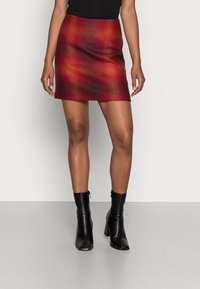 Nainen, joka pukeutuu punaisiin ja oransseihin ruutuhin minihaalariin, mustaan hihattomaan paitaan ja mustiin korkokenkisiin nilkkasaappaisiin, seisoo harmaalla lattialla.