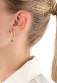 Guldörhänge med en tårformad grön ädelstenhängare, med slät yta och glänsande finish, bärs på vänster öra.