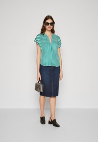 Stripet grønn og hvit kortermet bluse med V-hals, kombinert med en mørk denim blyantskjørt. Modellen holder en sølvveske og har på seg svarte sko.