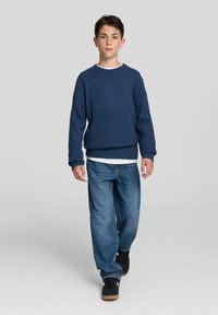 Granatowy sweter z teksturowanej dzianiny z białym kołnierzykiem, w połączeniu z jasnoniebieskimi spodniami dżinsowymi o luźnym kroju i czarnymi trampkami z brązową podeszwą.