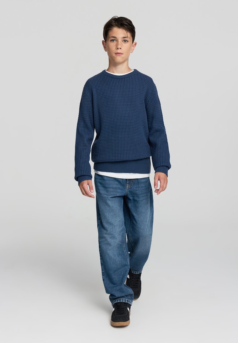 Granatowy sweter z teksturowanej dzianiny z białym kołnierzykiem, w połączeniu z jasnoniebieskimi spodniami dżinsowymi o luźnym kroju i czarnymi trampkami z brązową podeszwą.