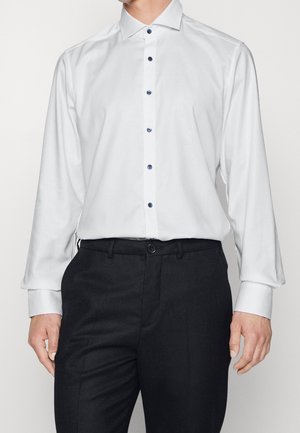 Mies, jolla on valkoinen pitkähihainen nappipaita, jossa on tummat napit, puettuna mustiin juhlahousuihin, seisoo yksinkertaisen taustan edessä.