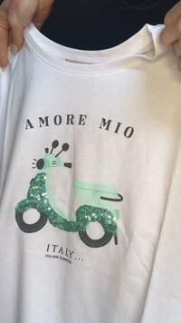 Bela bomba majica iz bombaža z grafiko zelene skuterja okrašeno s turkiznimi bleščicami. Besedilo pravi "AMORE MIO ITALIJA... ITALIJANSKA POLETJA."