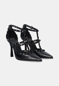 Zapatos de tacón alto de cuero negro con punta afilada, tiras transparentes y detalles con tachuelas en forma de pirámide. Presentan un tacón de aguja delgado.