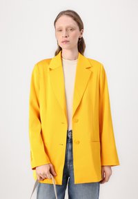 Lyse gul blazer med en strukturert design, doble knapper og en litt oversized passform, parret med en nøytral ribbestrikket topp og denim jeans.