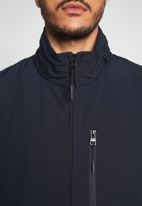 Granatowa kurtka z wysokim kołnierzem, zapinana z przodu na zamek błyskawiczny, z boczną kieszenią na zamek, wykonana z gładkiego, teksturowanego materiału.