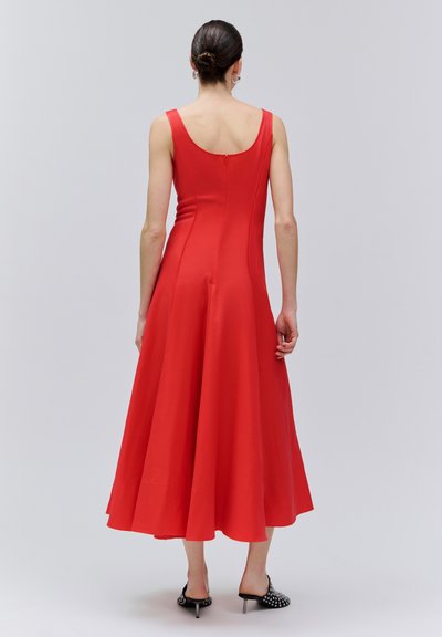 Femme aux cheveux noirs coiffés en chignon, portant une robe midi rouge sans manches et des mules à talons hauts noires cloutées, debout de dos sur un fond uni.