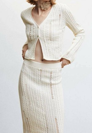 Mujer con un conjunto de punto cremoso de dos piezas, que incluye un cárdigan corto abotonado y una falda a juego de talle alto con patrones de cable vertical.