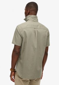 Camisa de manga corta de color verde oliva claro, hecha de una tela texturizada, que presenta un cuello contrario, pliegue en la espalda y sutilezas en los detalles de costura.