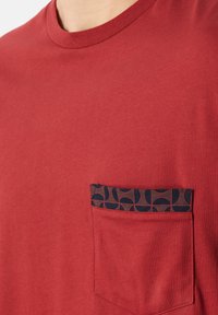 Rotes Baumwoll-T-Shirt mit rundem Ausschnitt und einer Tasche, die ein geometrisches Muster in Blau und Schwarz aufweist. Weiche Textur.