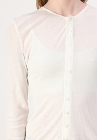 Langermet cardigan i kremfarge laget av lett stoff, med knappelukking foran og myk tekstur med en jevn overflate.