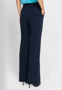 Granatowe spodnie z szerokimi nogawkami, wykonane z gładkiego materiału, z bocznymi kieszeniami i wysokim stanem. Nogawki opadają do podłogi, co podkreśla ich swobodny krój.