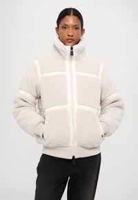 Fleece takki vaaleassa beigingissä, jossa on korkea kaulus, etuvetoketju ja kontrastivalkoiset paneelit. Varustettu sivutaskuilla ja ribbaatuilla hihansuilla.