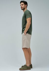 Camiseta de algodón verde oliva, pantalones cortos color beige claro y zapatillas verdes oliva. La vista lateral muestra un ajuste relajado y detalles de diseño casual.