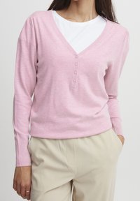 Suéter de punto de manga larga color rosa claro con escote en V, detalle de botones y puños acanalados, llevado sobre una camiseta blanca de cuello redondo.