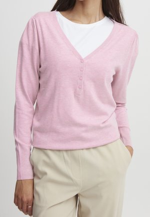 Lyserød langærmet pullover-trøje med V-hals, knapdetalje og ribstrikkede manchetter, bæret over en hvid crewneck-skjorte.