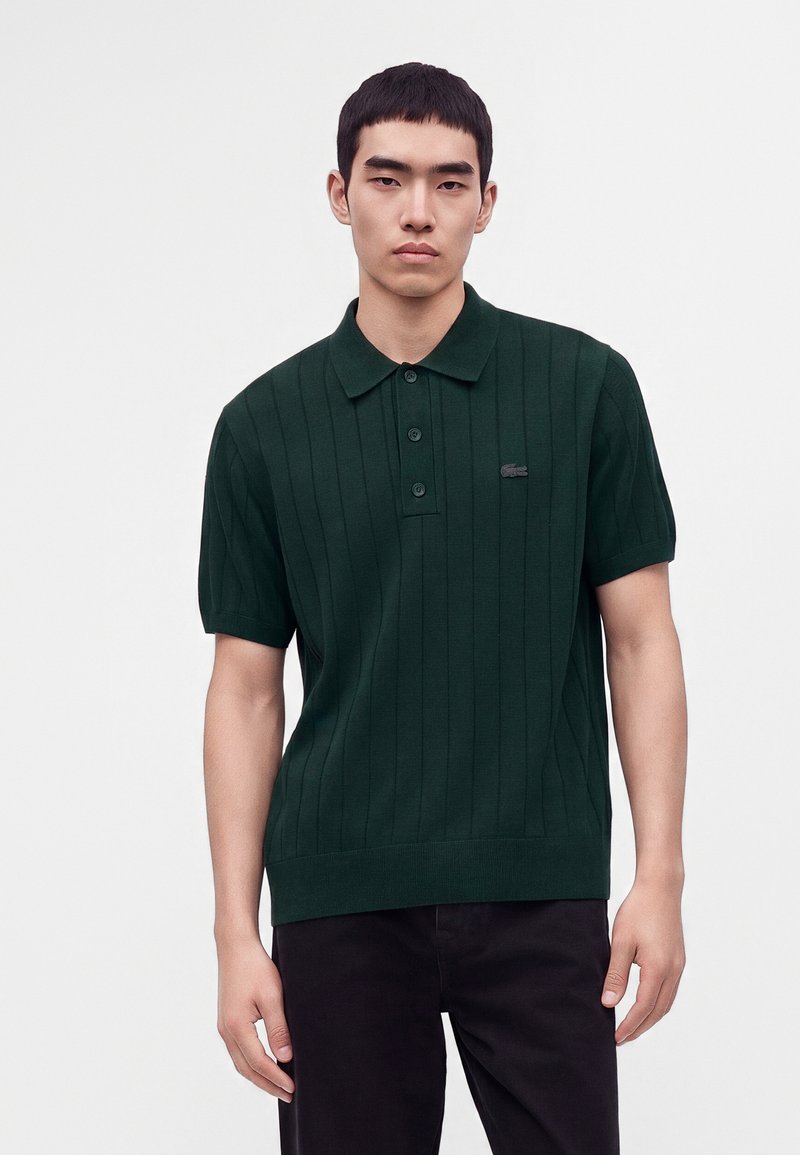 Jeune homme portant un polo vert foncé à manches courtes avec côte verticale et un pantalon noir, debout devant un fond clair uni.