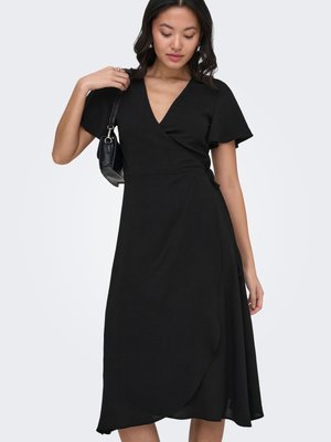 Nő fekete, rövid ujjú fodros felsővel rendelkező átlapolt ruhában, vállán kis fekete kézitáskával, egyszínű háttér előtt állva.