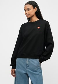 Svart genser med lange ermer, rund hals, ribbede mansjetter, med et lite rødt hjerte-logo på brystet, sammen med lyseblå jeans.