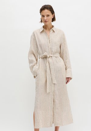 Frau, die ein langes, beige-weiß gestreiftes Hemdblusenkleid mit Gürtel, langen Ärmeln und Kragen trägt, vor einfachem weißem Hintergrund stehend.