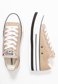 Béžové plátěné nízké tenisky Converse s bílými gumovými podrážkami, černým pruhovaným detailem, bílými tkaničkami a kovovými očky na bílém pozadí.
