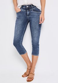 Jean capri en denim bleu foncé avec une taille mi-haute, présentant des poches avant et des ourlets retroussés. Associé à des sandales brunes à lanières.