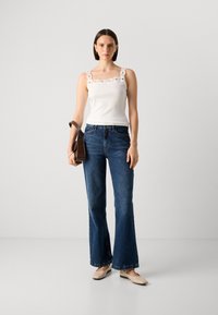 White tank top with lace trim, blue flared jeans, and beige flats. Model holding a brown shoulder bag. Plain gray background.