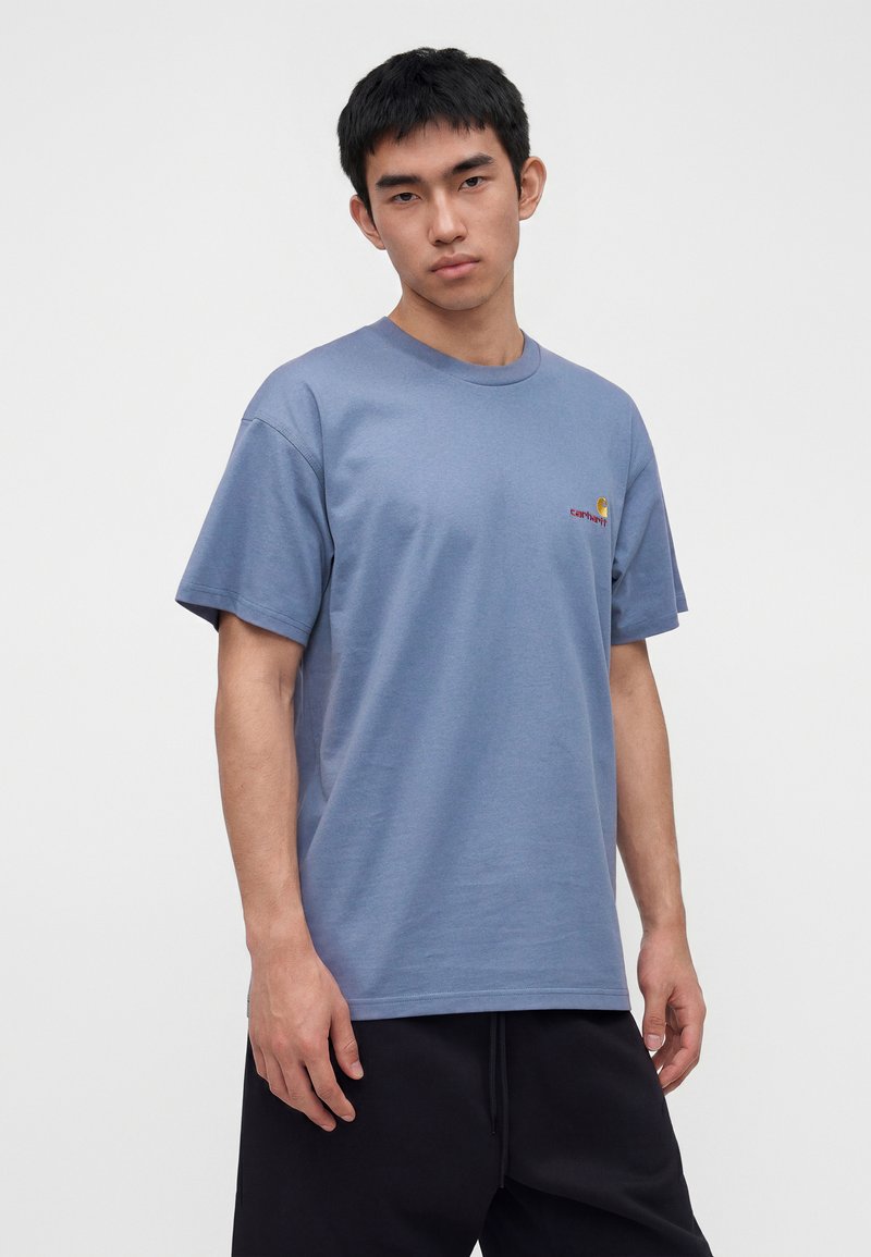 Young man wearing a blue Carhartt t-shirt and black pants, standing against a plain light background, looking at the camera.