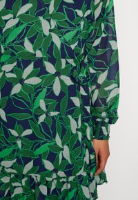 Sukienka w kwiaty z przezroczystego materiału, z granatowym tłem, zielonymi liśćmi i białymi kwiatami, z falbaniastym dołem i marszczonymi mankietami.