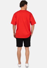 T-shirt rossa in cotone con maniche corte, vestibilità ampia, orlo posteriore leggermente più lungo, abbinata a pantaloni corti neri e sneakers bianche.