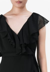 Frau in einem schwarzen Kleid mit transparenten, rüschierten Kurzärmeln und einem tiefen V-Ausschnitt, vom Hals bis zur Taille vor einem weißen Hintergrund zu sehen.