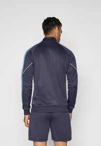 Chaqueta deportiva en color marino con manga larga, que presenta acentos en azul claro en los hombros, textura suave y un cuello alto.