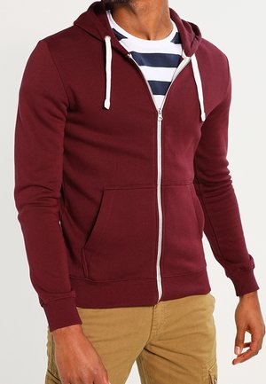 Uomo che indossa una felpa con cappuccio bordeaux con zip e cordini bianchi sopra una camicia a righe blu navy e bianca e pantaloni beige.