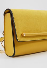 Borsa a tracolla in suede e pelle gialla dal design elegante, con una barra di accento dorato e piping nero lungo i bordi. Cinghia regolabile inclusa.
