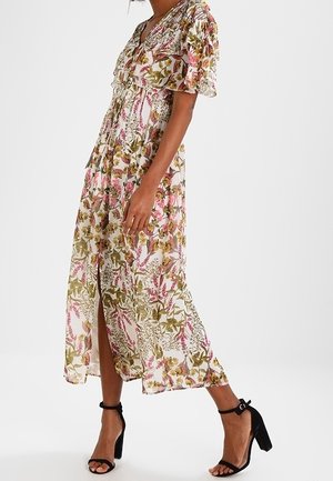 Kvinde iført en blomstrede midi-kjole med flagrende ærmer og en frontslids, kombineret med sorte ankelremshæle.