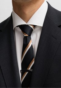 Cravatta a strisce nera e dorata con una texture liscia, caratterizzata da linee diagonali e una barretta metallica, indossata con una camicia bianca e un giaccone scuro.