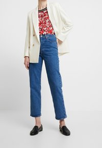Persoon in een witte blazer, met een rode bloemen-top, blauwe high-waisted jeans en zwarte instappers, staande met één hand op de heup.