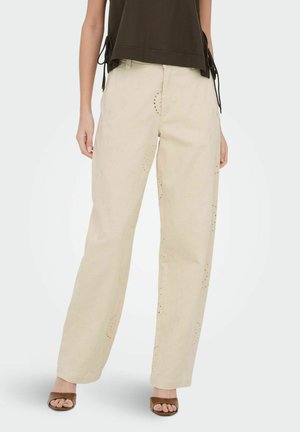 Femme portant un pantalon beige à jambes larges avec des découpes florales subtiles, associé à des talons ouverts marron et un top noir sans manches.