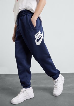 Osoba w granatowych spodniach dresowych Nike z białym logo i białych butach Nike, stojąca na szarej podłodze na tle jasnej, gładkiej ściany.