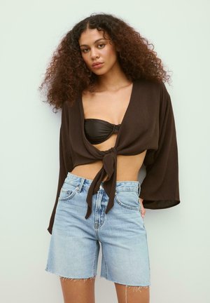 Frau mit lockigem Haar trägt ein schwarzes Bandeau, ein locker gebundenes dunkelbraunes Hemd und hellblaue Jeansshorts mit ausgefransten Säumen, steht vor einer blassen Wand.