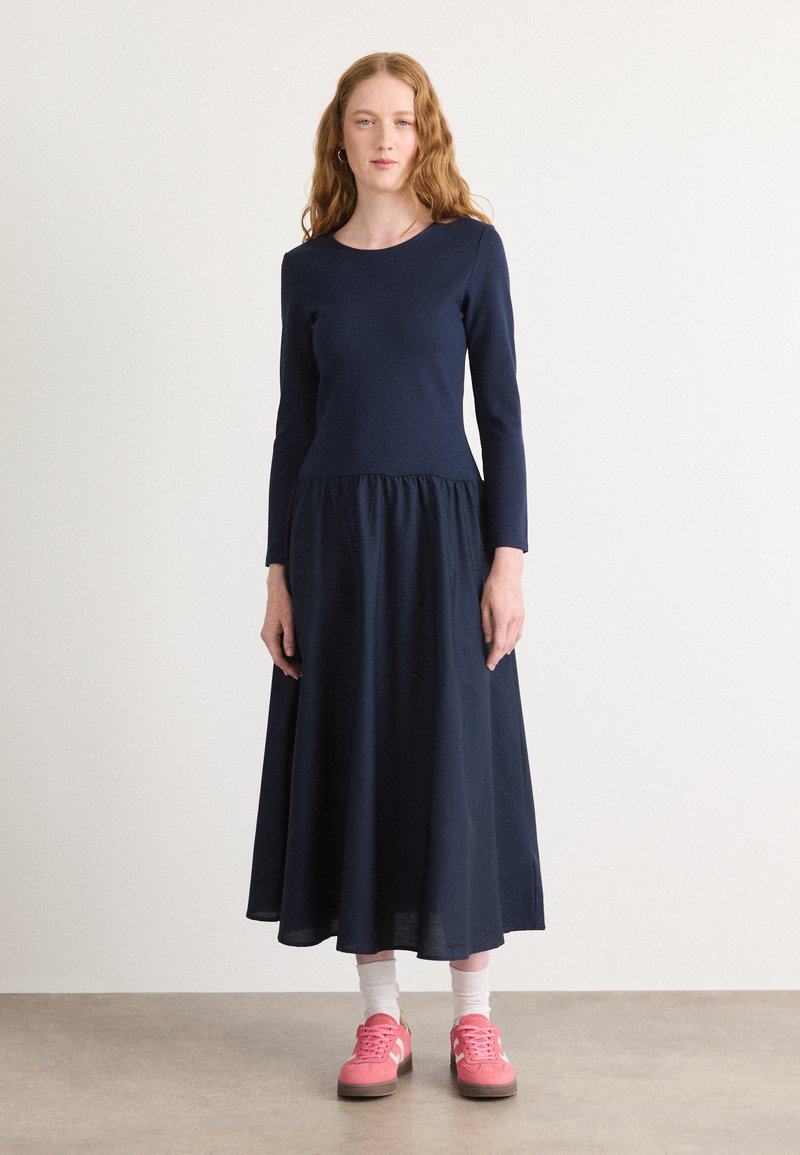 Abito lungo a maniche di colore blu navy con vita arricciata e gonna svasata. Indossato con calzature rosa e calze bianche, su uno sfondo semplice.