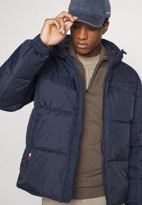 Navy pufferjakke laget av quiltet stoff, med frontglidelås, ståkrage og merkepatch på brystet. Myk innerside synlig.