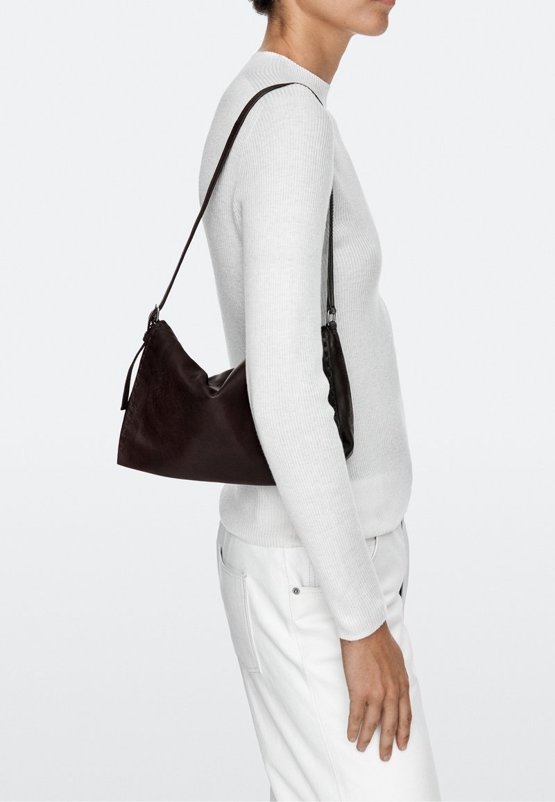 Woman wearing a white ribbed sweater and white pants carrying a black leather shoulder bag with a thin strap on her right shoulder.