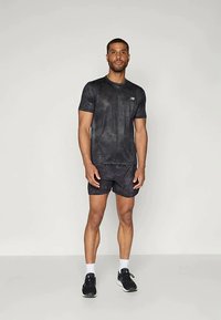 Mees seisab seljas must New Balance'i sportlik T-särk, sobivad lühikesed püksid, mustad spordijalanõud ja valged sokid, taustal lihtne heledat värvi taust.