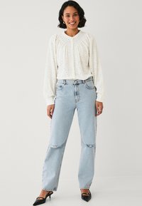 Blusa bianca in cotone con ricami e maniche lunghe, dal collo rotondo, abbinata a jeans strappati a vita alta di colore blu chiaro e tacchi neri a punta.