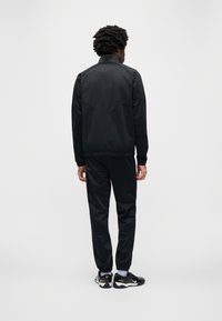 Μαύρο αθλητικό μπουφάν με όρθιο γιακά, λεία υφή και λεπτομέρεια πίσω ώμου; συνδυάζεται με παντελόνι στο ίδιο χρώμα και μαύρα αθλητικά παπούτσια.