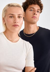 Divas modeļi stāv blakus. Kreisais valkā pieguļošu krēmkrāsas ribotu T-kreklu un zelta kaklarotu, bet labais valkā tumši zilu T-kreklu ar līdzīgu kaklarotu.